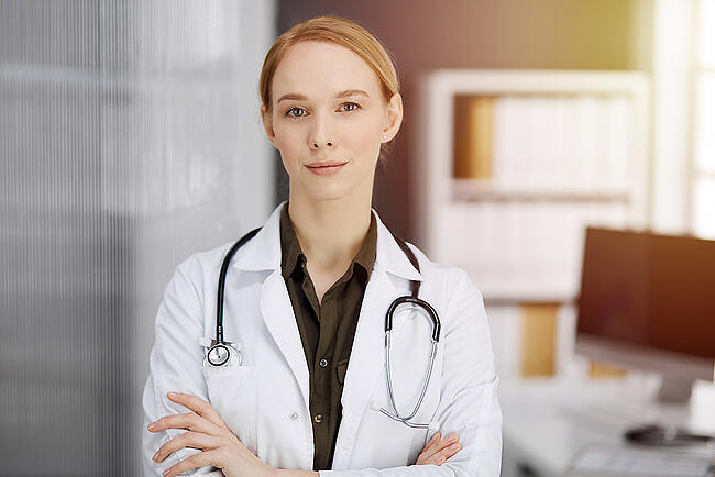 Nurse scheduler wearing a brown shirt with a white coat over it  standing with her arms crossed.
