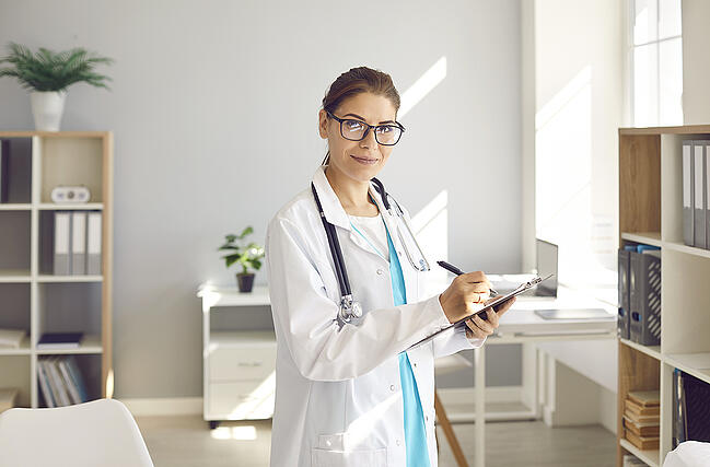 Nurse manager wearing glasses holding a clipboard and writing on it with a black pen.