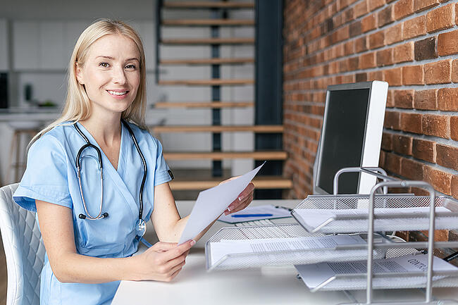 Nurse manager wear light blue scrubs sitting at a desk looking through paperwork.