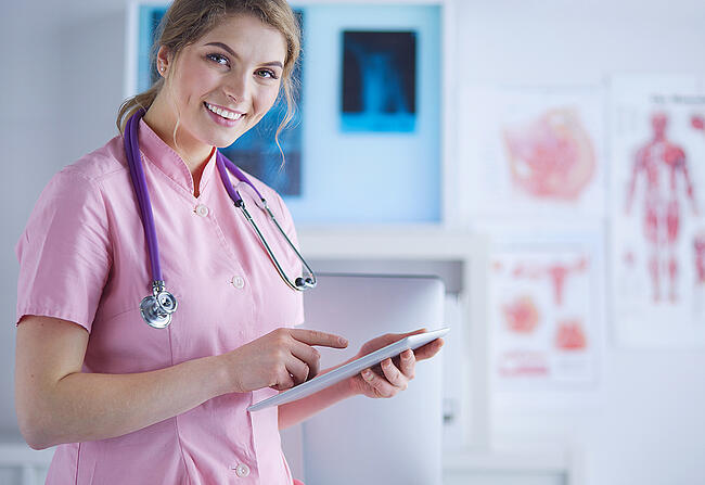 Female nurse wearing pink scrubs smiling at the camera while holding a tablet.